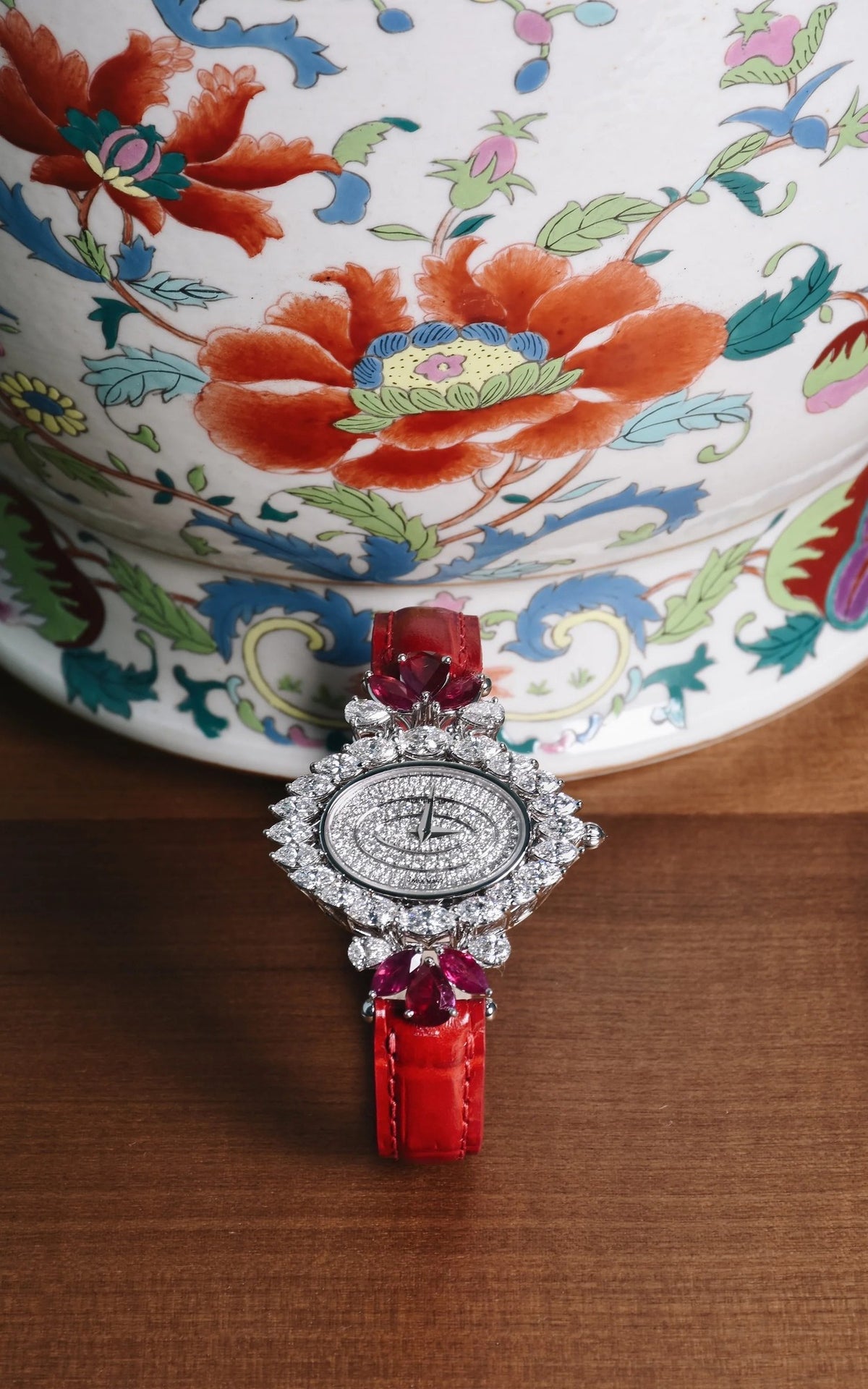 An oval diamond watch with rubies, on a red strap, standing before a painted porcelain vase.