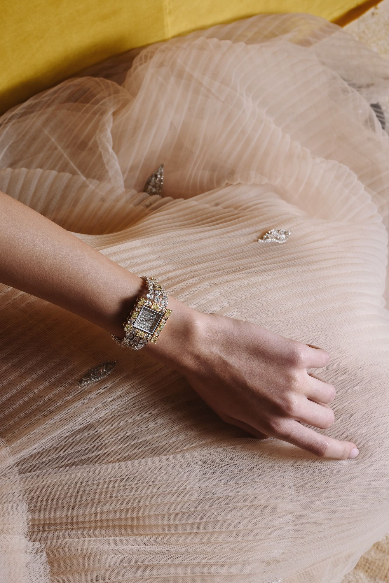 A diamond-set watch worn at the wrist, a hand resting on pleated tulle
